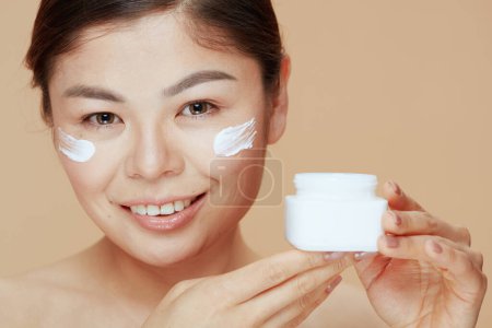 Foto de Retrato de mujer asiática moderna con frasco de crema facial y crema facial en la cara contra fondo beige. - Imagen libre de derechos
