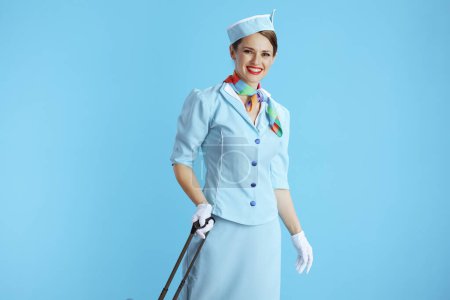 Téléchargez les photos : Heureuse élégante hôtesse de l'air femme sur fond bleu en uniforme bleu avec sac de voyage. - en image libre de droit
