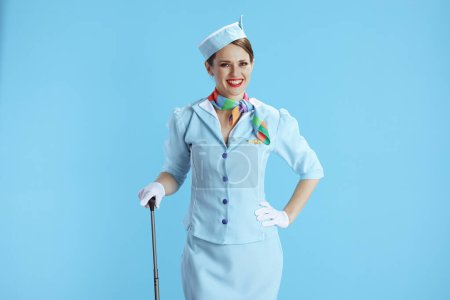 Téléchargez les photos : Heureuse hôtesse de l'air féminine élégante isolée sur fond bleu en uniforme bleu avec sac de voyage. - en image libre de droit