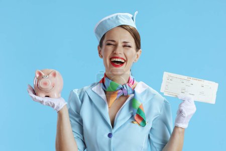 Photo for Happy modern female stewardess against blue background in blue uniform with flight tickets and piggy bank. - Royalty Free Image