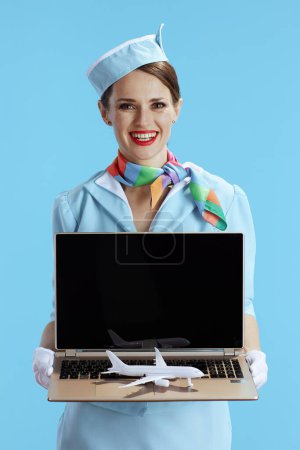 Photo for Happy stylish air hostess woman against blue background in blue uniform with a little airplane showing laptop blank screen. - Royalty Free Image