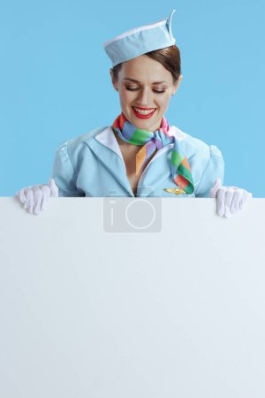 Téléchargez les photos : Joyeuse hôtesse de l'air élégante sur fond bleu uniforme bleu montrant tableau blanc. - en image libre de droit