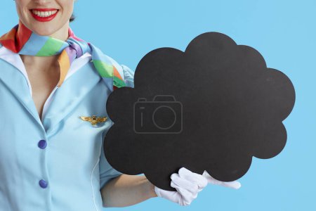 Téléchargez les photos : Gros plan sur une hôtesse de l'air souriante sur fond bleu en uniforme bleu montrant un panneau de forme de nuage vierge. - en image libre de droit