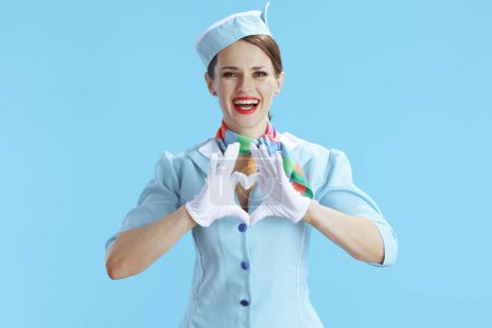 Téléchargez les photos : Hôtesse de l'air féminine moderne souriante sur fond bleu en uniforme bleu montrant les mains en forme de coeur. - en image libre de droit