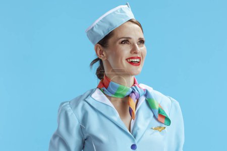 Foto de Sonriente elegante azafata de aire femenino sobre fondo azul en uniforme azul mirando a la distancia. - Imagen libre de derechos