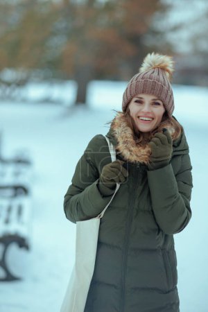 Photo for Happy modern middle aged woman in green coat and brown hat outdoors in the city park in winter with mittens and beanie hat. - Royalty Free Image