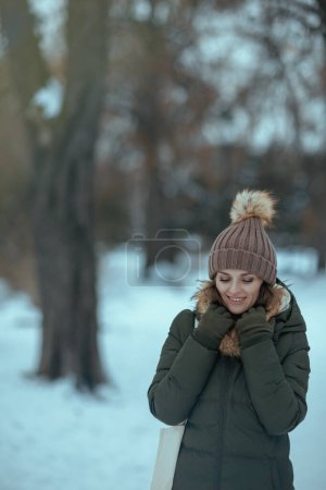 Photo for Smiling modern 40 years old woman in green coat and brown hat outdoors in the city park in winter with mittens and beanie hat. - Royalty Free Image