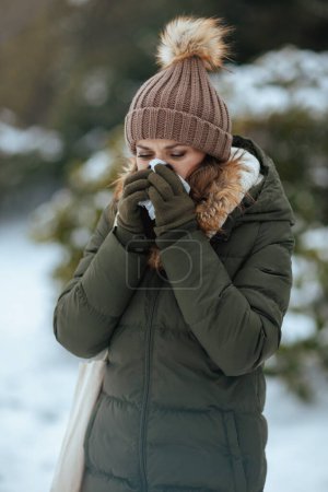Photo for Sad modern female in green coat and brown hat outdoors in the city park in winter with mittens, napkin blowing nose and beanie hat. - Royalty Free Image