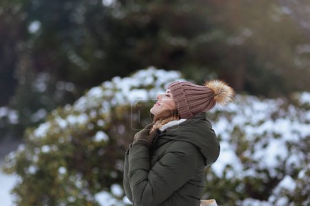 Photo for Smiling modern woman in green coat and brown hat outdoors in the city park in winter with beanie hat near snowy branches. - Royalty Free Image