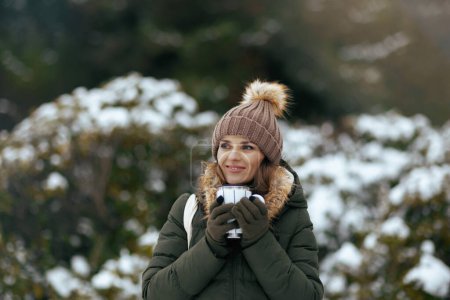 Foto de Mujer moderna feliz en abrigo verde y sombrero marrón al aire libre en el parque de la ciudad en invierno con manoplas, taza de café y gorro sombrero. - Imagen libre de derechos