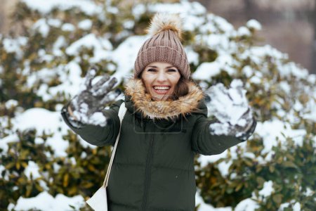 Foto de Feliz mujer moderna de 40 años en abrigo verde y sombrero marrón al aire libre en el parque de la ciudad en invierno con manoplas nevadas y gorro cerca de ramas nevadas. - Imagen libre de derechos