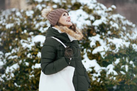Foto de Sonriente mujer moderna de mediana edad en abrigo verde y sombrero marrón al aire libre en el parque de la ciudad en invierno con manoplas y gorro sombrero cerca de ramas nevadas. - Imagen libre de derechos