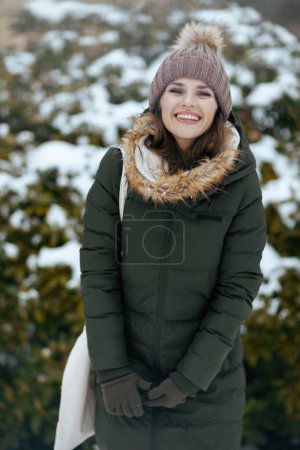 Foto de Sonriente mujer moderna en abrigo verde y sombrero marrón al aire libre en el parque de la ciudad en invierno con manoplas y gorro sombrero cerca de ramas nevadas. - Imagen libre de derechos