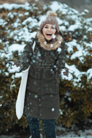 Téléchargez les photos : Femme moderne souriante en manteau vert et chapeau marron à l'extérieur dans le parc de la ville en hiver avec des mitaines et un bonnet jouant avec la neige près des branches enneigées. - en image libre de droit