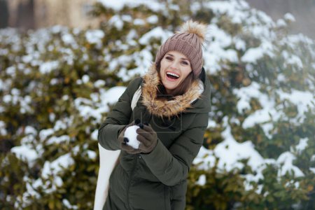 Foto de Sonriente mujer moderna en abrigo verde y sombrero marrón al aire libre en el parque de la ciudad en invierno con manoplas y gorro lanzando bolas de nieve cerca de ramas nevadas. - Imagen libre de derechos