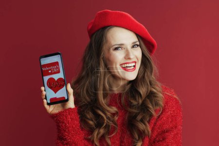 Téléchargez les photos : Joyeux Valentin. Femme moderne souriante avec de longs cheveux ondulés en pull rouge et béret, smartphone et application Valentine. - en image libre de droit
