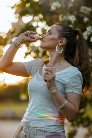 Foto de Hora de verano. mujer de moda en camisa blanca usando aerosol nasal cerca del árbol floreciente. - Imagen libre de derechos