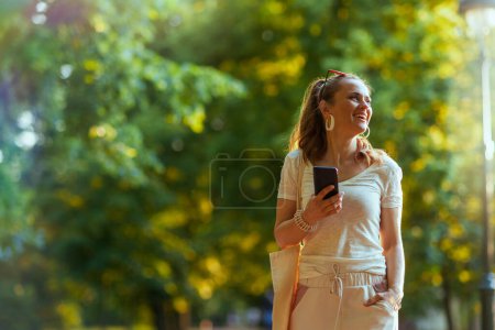 Foto de Hora de verano. feliz mujer moderna de 40 años de edad en camisa blanca con bolsa de mano utilizando aplicaciones de teléfonos inteligentes y caminando en el parque de la ciudad. - Imagen libre de derechos