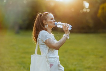 Foto de Hora de verano. mujer de moda de 40 años en camisa blanca con bolsa de mano y botella de agua en el prado al aire libre. - Imagen libre de derechos