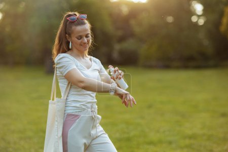 Foto de Hora de verano. mujer de moda feliz en camisa blanca con bolsa de mano usando pulverización de ácaros en el prado exterior en la naturaleza. - Imagen libre de derechos