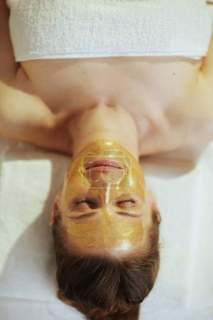 Photo for Healthcare time. Upper view of relaxed modern woman in massage cabinet with golden cosmetic mask on face laying on massage table. - Royalty Free Image