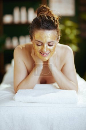 Téléchargez les photos : Temps de santé. heureuse femme moderne dans le salon de spa avec masque cosmétique doré sur la pose du visage sur la table de massage. - en image libre de droit
