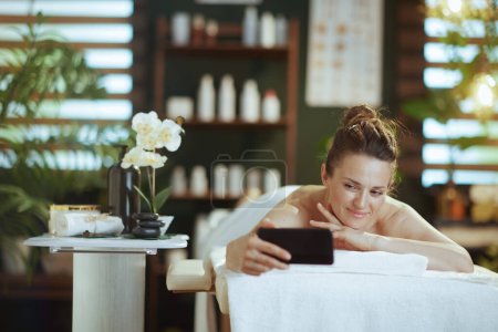 Téléchargez les photos : Temps de santé. souriant moderne femme d'âge moyen dans l'armoire de massage prendre selfie et la pose sur la table de massage. - en image libre de droit