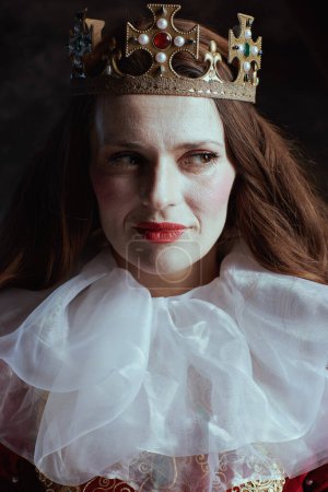 Téléchargez les photos : Reine médiévale coûteuse en robe rouge avec col blanc et couronne sur fond gris foncé. - en image libre de droit