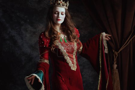 Photo for Medieval queen in red dress with white makeup and crown on dark gray background. - Royalty Free Image