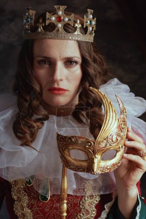 Téléchargez les photos : Reine médiévale en robe rouge avec masque vénitien, col blanc et couronne sur fond gris foncé. - en image libre de droit