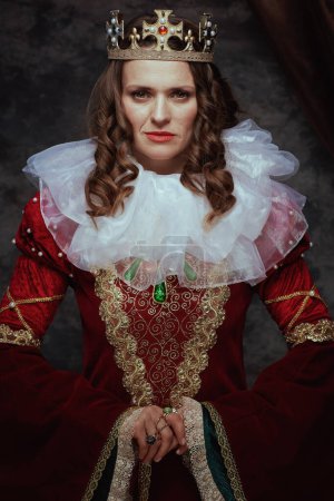 Photo for Medieval queen in red dress with white collar and crown on dark gray background. - Royalty Free Image