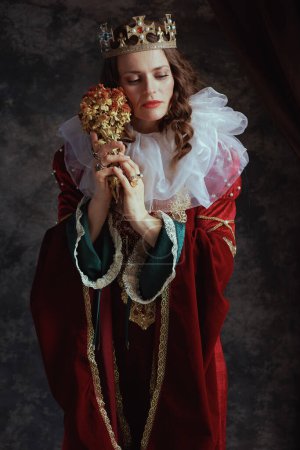 Foto de Reina medieval pensativa en vestido rojo con flor seca, cuello blanco y corona sobre fondo gris oscuro. - Imagen libre de derechos