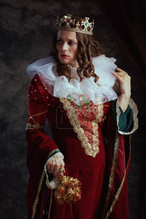Téléchargez les photos : Reine médiévale stressée en robe rouge avec fleur séchée, col blanc et couronne sur fond gris foncé. - en image libre de droit