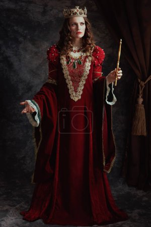 Foto de Retrato de longitud completa de la reina medieval en vestido rojo con varita y corona sobre fondo gris oscuro. - Imagen libre de derechos