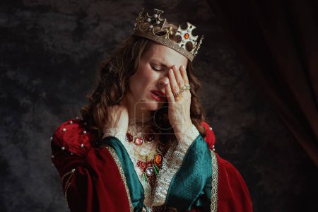 Foto de Reina medieval acentuada en vestido rojo con corona que tiene dolor de cuello sobre fondo gris oscuro. - Imagen libre de derechos
