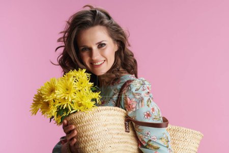 Foto de Retrato de mujer elegante y sonriente de mediana edad en vestido floral con flores de crisantemos amarillos y bolsa de paja aislada sobre fondo rosa. - Imagen libre de derechos