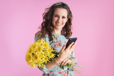 Photo for Portrait of happy trendy woman with long wavy brunette hair with yellow chrysanthemums flowers sending text message using smartphone isolated on pink. - Royalty Free Image