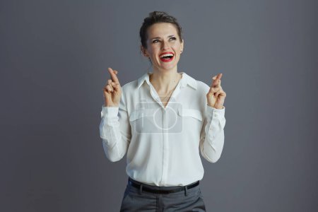 Téléchargez les photos : Souriant moderne d'âge moyen propriétaire de petite entreprise femme en chemisier blanc avec les doigts croisés isolé sur gris. - en image libre de droit