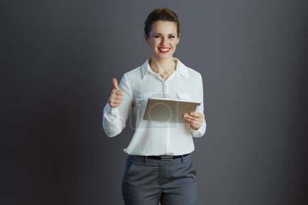 Téléchargez les photos : Souriant jeune femme travailleur en chemisier blanc à l'aide de la tablette PC montrant pouces isolés sur gris. - en image libre de droit