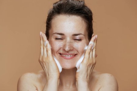 Foto de Mujer de mediana edad feliz con espuma facial limpiador lavando la cara contra el fondo beige. - Imagen libre de derechos