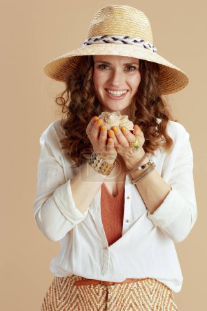 Photo for Beach vacation. happy stylish 40 years old housewife in white blouse and shorts against beige background with seashell and summer hat. - Royalty Free Image