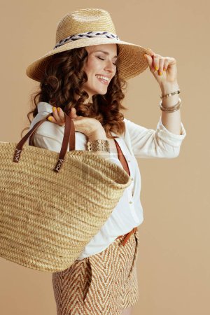 Foto de Vacaciones en playa. mujer elegante feliz en blusa blanca y pantalones cortos aislados en fondo beige con bolsa de paja y sombrero de paja. - Imagen libre de derechos