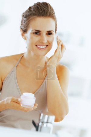 Foto de Sonriente mujer joven aplicando crema en el baño - Imagen libre de derechos