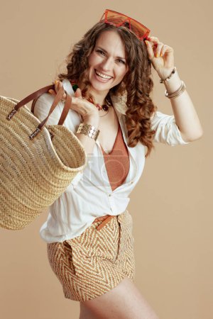 Foto de Vacaciones en playa. mujer moderna feliz en blusa blanca y pantalones cortos sobre fondo beige con bolsa de paja y gafas de sol. - Imagen libre de derechos