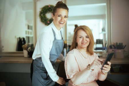 Téléchargez les photos : Femme de 40 ans coiffeuse dans un studio de coiffure moderne avec client avec smartphone. - en image libre de droit