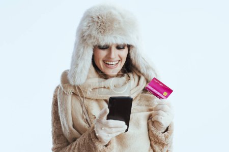 Foto de Mujer con estilo feliz en abrigo de invierno y sombrero de piel sobre fondo blanco en guantes blancos con tarjeta de crédito y teléfono inteligente. - Imagen libre de derechos