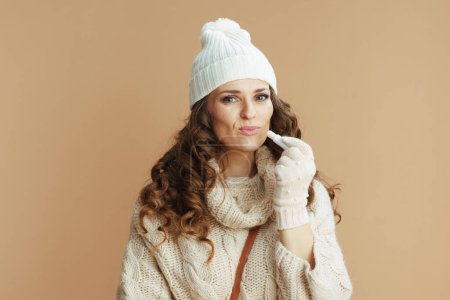 Téléchargez les photos : Bonjour l'hiver. femme élégante en pull beige, mitaines et chapeau avec rouge à lèvres hygiénique isolé sur beige. - en image libre de droit