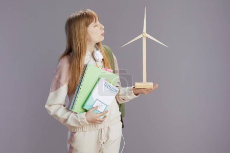 Photo for Trendy teenage girl in beige tracksuit with workbooks, headphones, windmill and textbook isolated on grey background. - Royalty Free Image