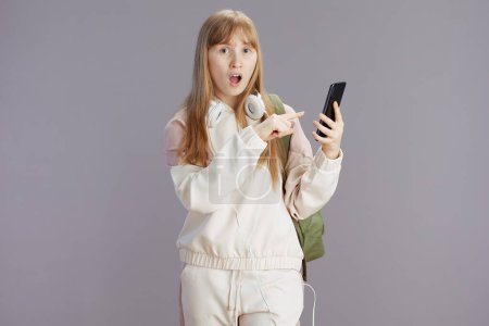 Foto de Sorprendida chica adolescente moderna en chándal beige con mochila y auriculares usando teléfono inteligente aislado en gris. - Imagen libre de derechos