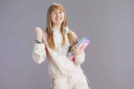 Photo for Portrait of smiling trendy teen girl in beige tracksuit with workbooks and headphones isolated on grey background rejoicing. - Royalty Free Image